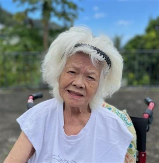An elderly woman with white hair smiles brightly while seated outdoors in a calm environment.