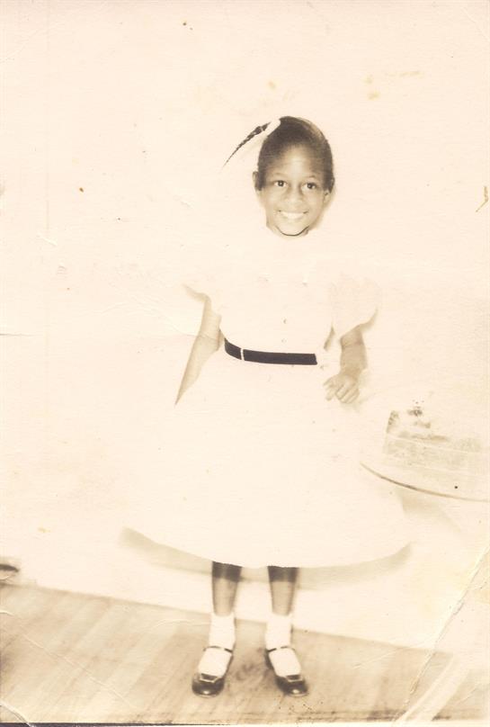 A young girl smiles while wearing a white dress with a black belt, posing cheerfully indoors.