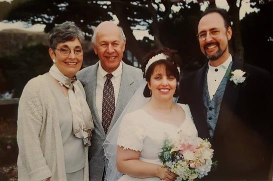 A bride stands with her parents and husband, all smiling warmly in a beautiful outdoor location.