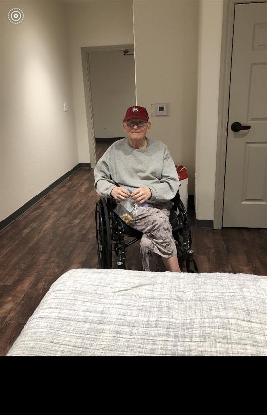 Elderly man in wheelchair seated inside a plain room, likely resting or waiting.