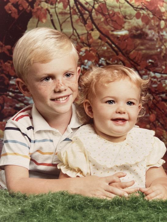 Two young siblings are seated on green grass, smiling happily against colorful autumn leaves.