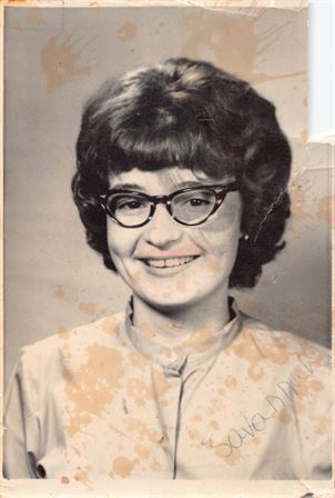 A cheerful woman with glasses poses, showing a friendly smile and classic hairstyle.