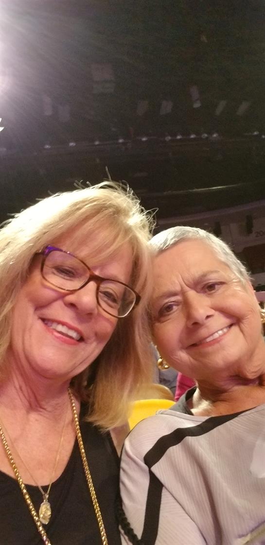 Two women share a warm smile while enjoying a lively event in a crowded hall.