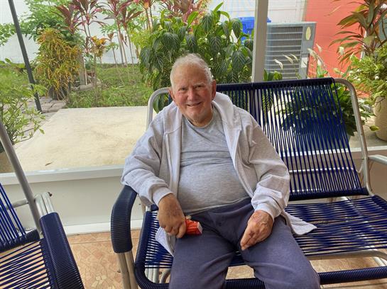 An elderly man relaxes on a bench surrounded by lush greenery in a warm outdoor setting.