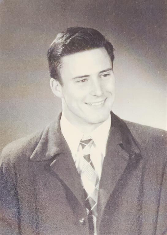 A young man stands confidently, dressed in a formal coat and tie, smiling warmly towards the camera.