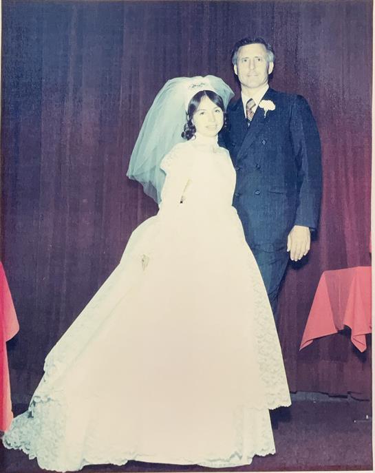 Bride in a white gown stands with her father in tuxedo at a reception hall with table settings.