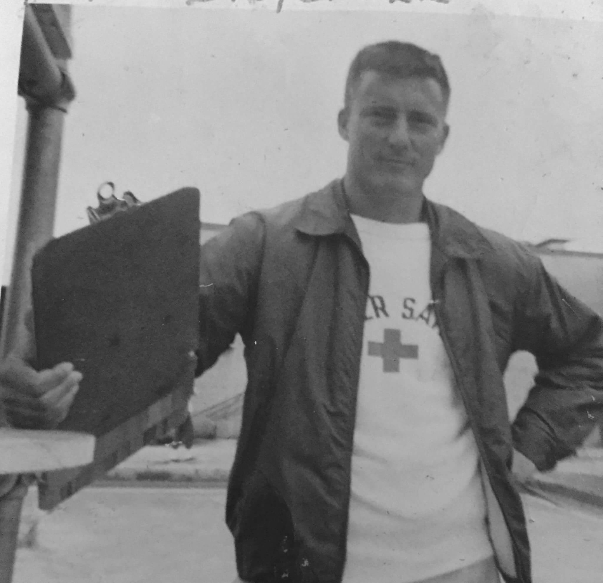 A man poses confidently on the deck of a naval vessel while holding a clipboard in the 1960s.