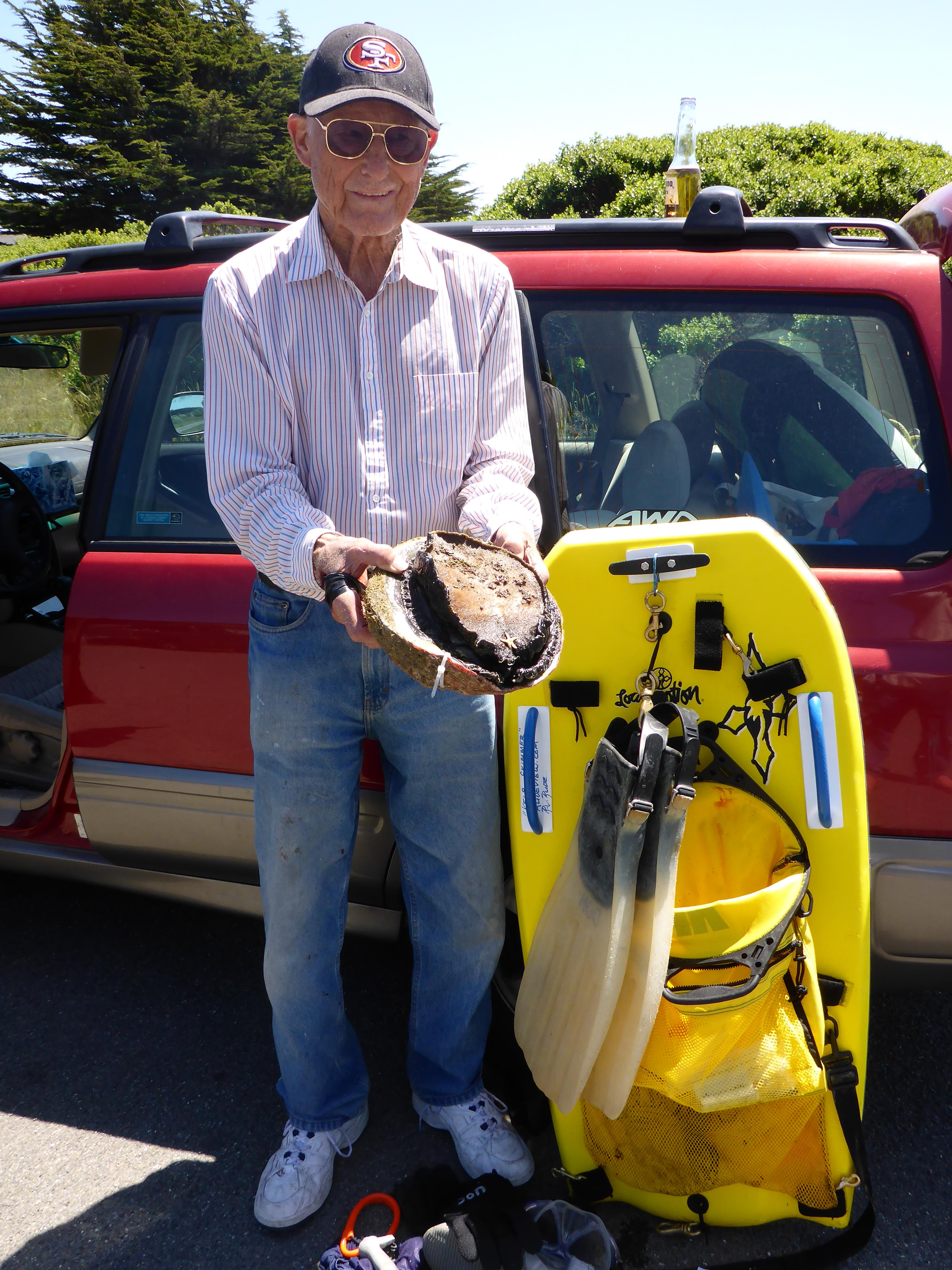 An elderly man smiles with a large shell by a bright yellow kayak at the coast.