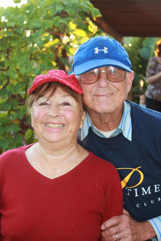 Two smiling adults share a warm embrace during a sunny day at a garden event.