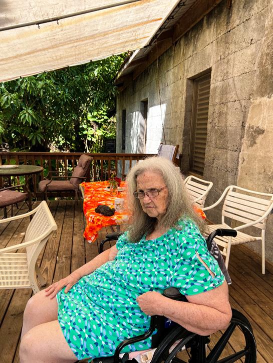 An elderly woman in a wheelchair sits on a deck, surrounded by furniture and greenery.