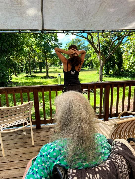 A woman is stretching outdoors on a deck while an older person sits quietly nearby, enjoying nature.