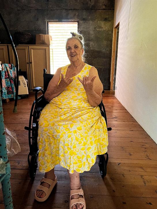 Elderly woman happily gestures from her wheelchair in a warm, inviting space with wooden floors.