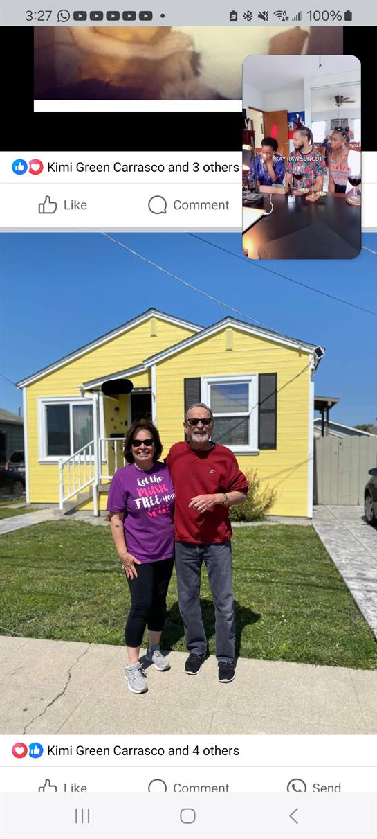 Two people stand in front of a cheerful yellow house on a clear day, smiling and relaxed.