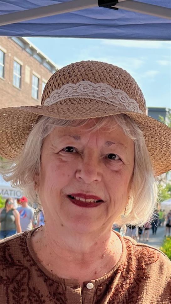 A cheerful woman with short gray hair wears a straw hat at a vibrant outdoor market.