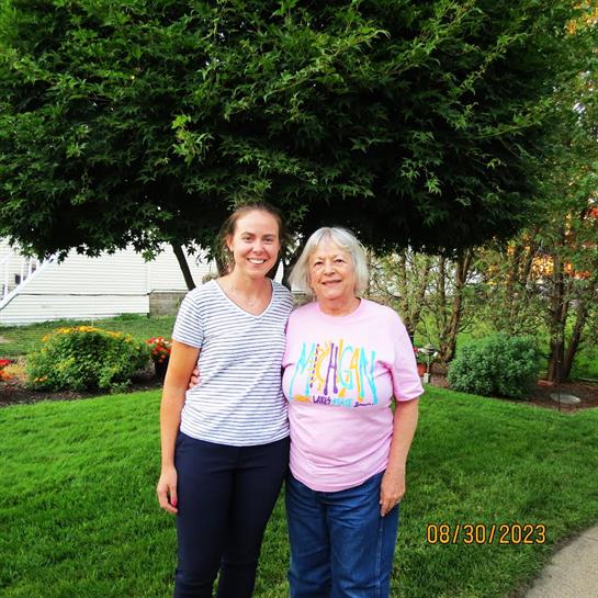 Two women stand together in a lush garden, both smiling happily in summer attire.