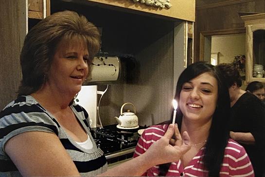 Two women joyfully prepare for a birthday celebration by lighting a candle in a kitchen.