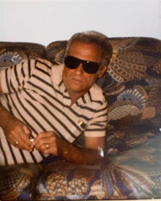 An older man in sunglasses lounges comfortably on a decorative couch while holding a lighter.