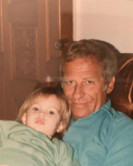 An older man smiles gently while holding a young child in his lap, both looking relaxed at home.