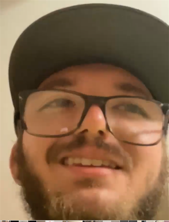 A young man with glasses and a beard smiles while in a casual indoor setting.