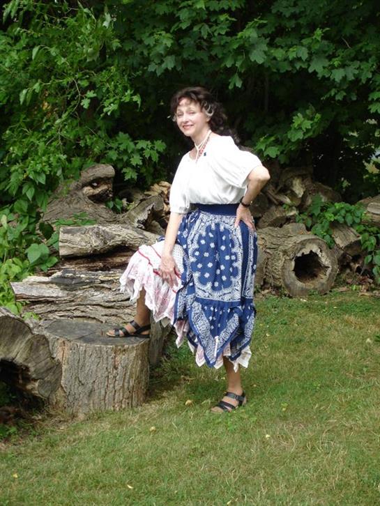 A woman in a white top and blue polka dot skirt poses playfully next to large garden logs.