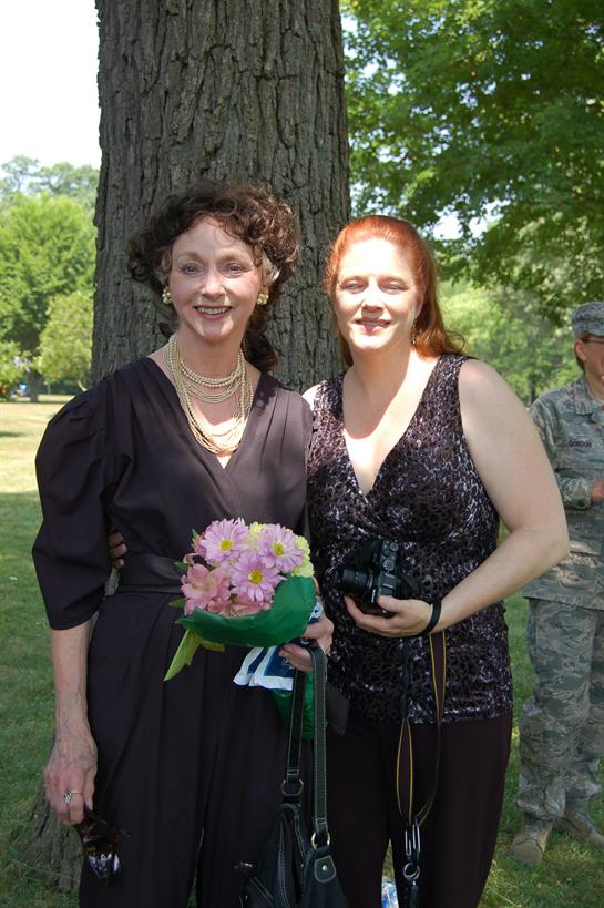 Two women smile together, surrounded by greenery, holding flowers and cameras at a joyful event.