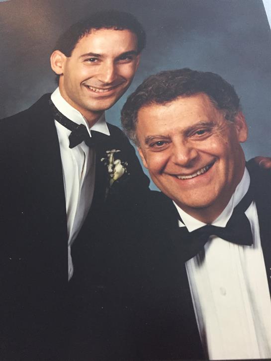 A father and son smile warmly at a special gathering, dressed in tuxedos and celebrating together.