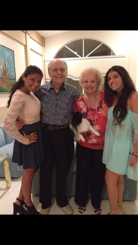 Four family members smile together while holding a small dog in a warm living room setting.