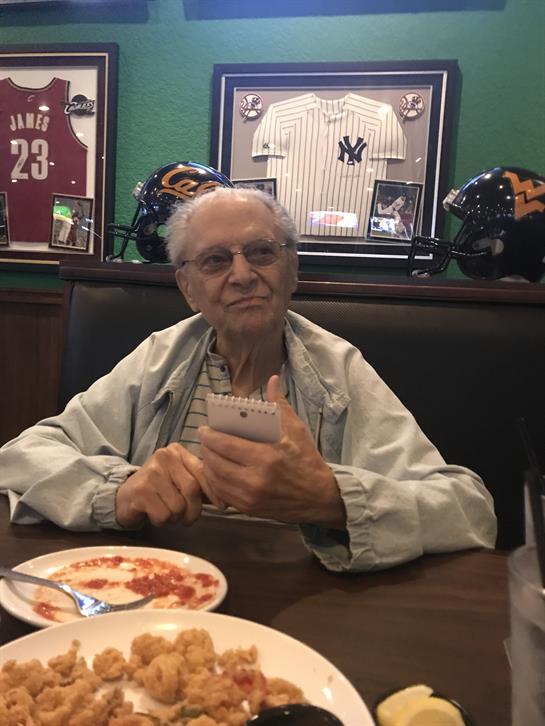 An elderly man sitting at a table with food, engaged with his mobile device in a lively atmosphere.