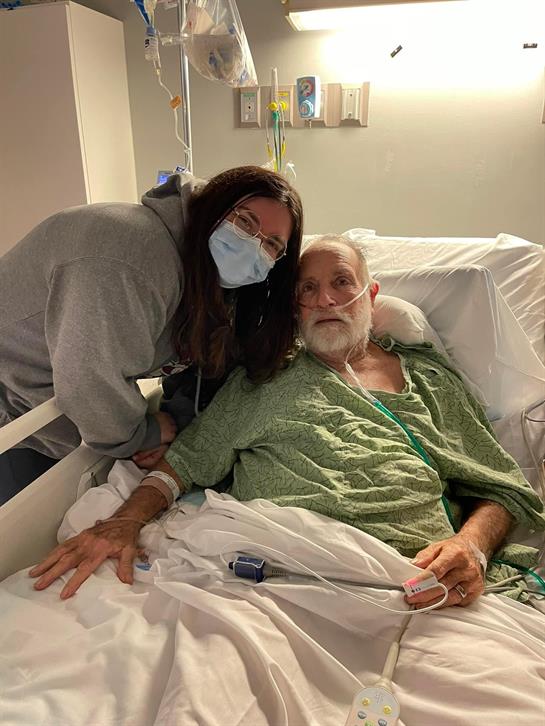 A woman in a mask smiles beside an elderly man in a hospital bed, showing support.