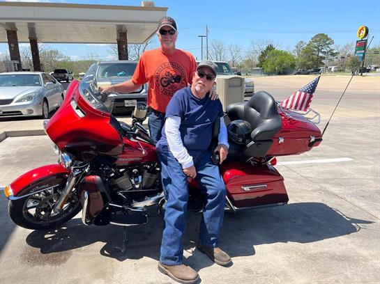 Two men are posing near a bright red motorcycle at a roadside location on a sunny day.