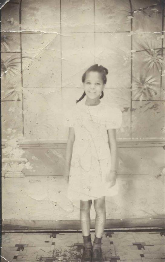 A young girl stands smiling in a vintage dress in front of an ornate floral pattern backdrop.