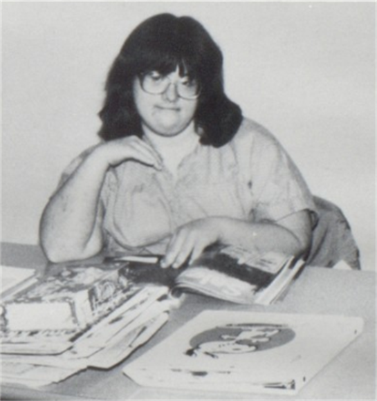 A woman with glasses sits at a desk filled with papers, appearing contemplative in a casual setting.