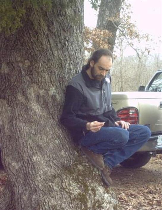 A man sits against a tree and takes notes while a truck is parked nearby in the woods.