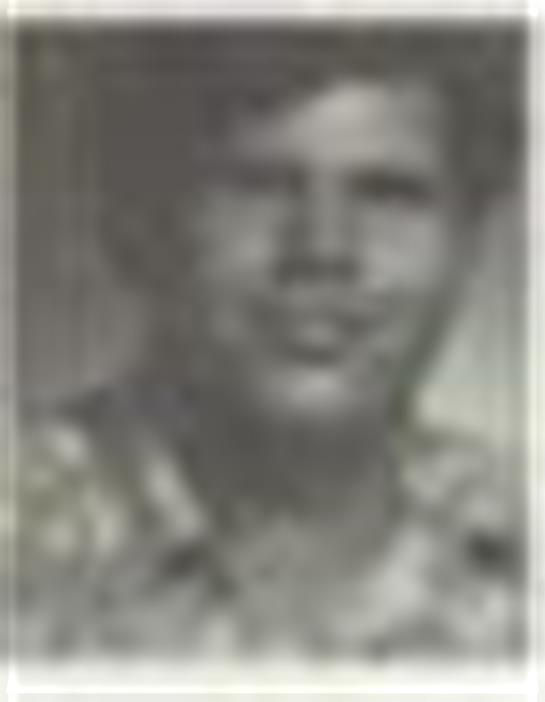 A man with a neutral expression wears a floral shirt and poses for a portrait in soft focus.