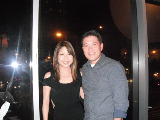 Two people smile and pose close together in front of large windows at night.