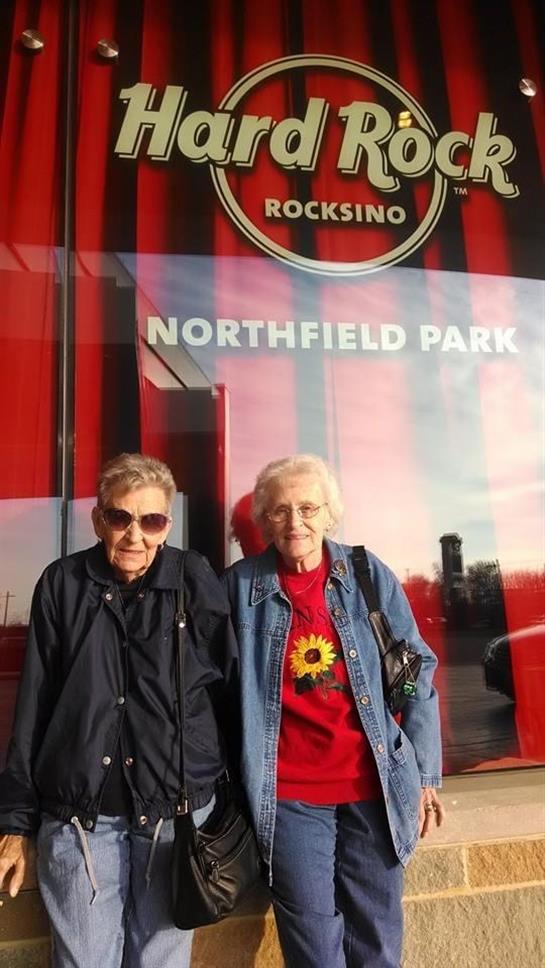 Two older women stand smiling outside a casino, wearing casual clothing and sunglasses.