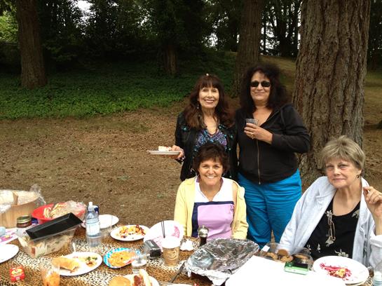 Four women gather around a picnic table filled with food and drinks, smiling and chatting.