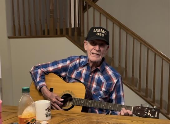 An elderly man plays guitar and sings at home, wearing a military cap and checked shirt.