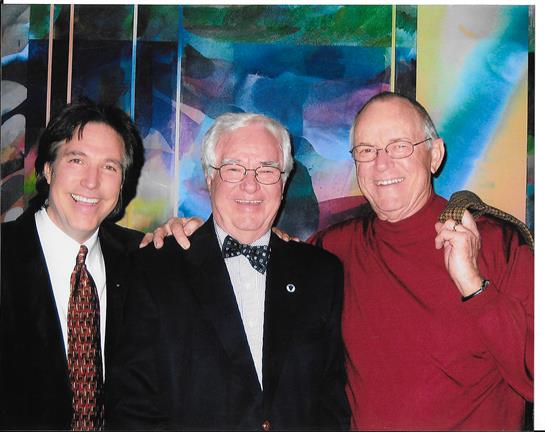 Three men smile and pose together at a social gathering with a vibrant art backdrop.