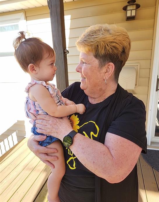 A grandmother smiles warmly while holding her granddaughter on a wooden porch.