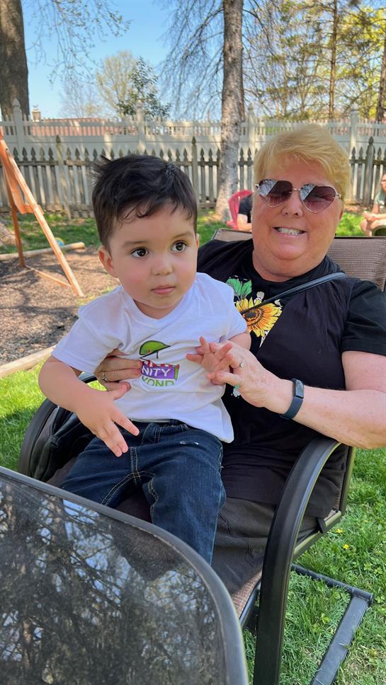 A young boy sits on a woman's lap in a park, both smiling happily in the sun.