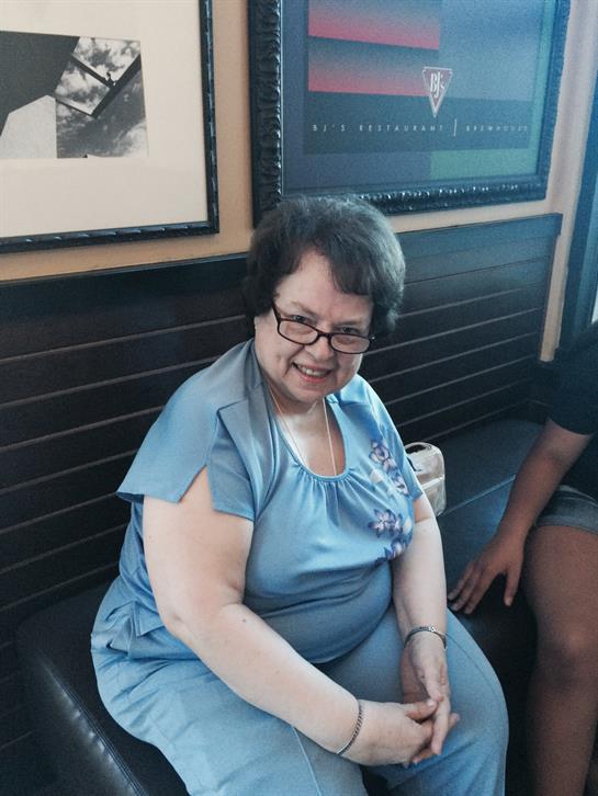 An elderly woman wearing a blue outfit sits on a bench in a relaxed cafe atmosphere, smiling warmly.