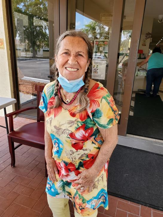 A joyful elderly woman with braids stands outside a shop wearing a floral dress and mask.