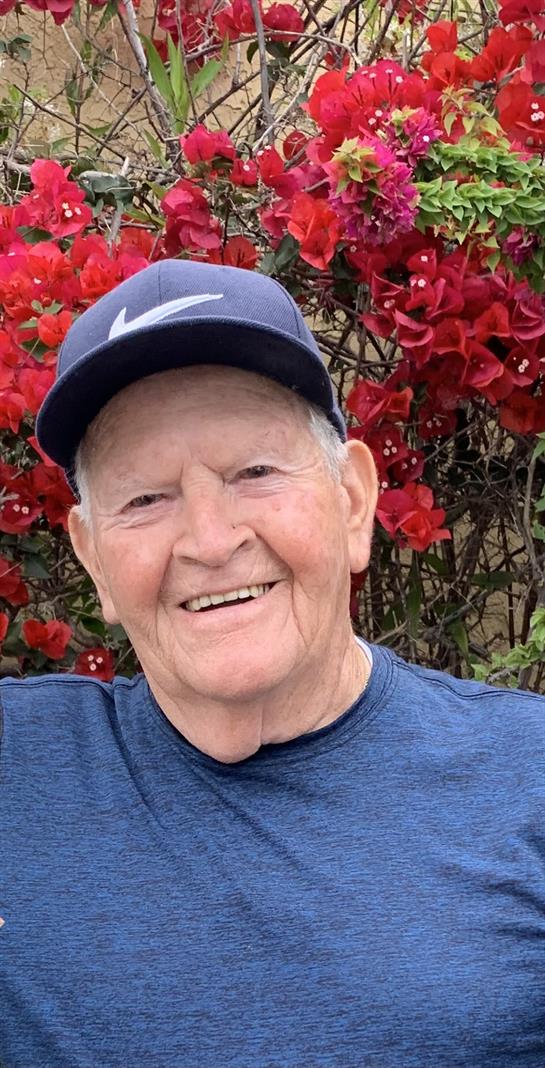Elderly man with a warm smile enjoys a sunny day among blooming red flowers in a garden.