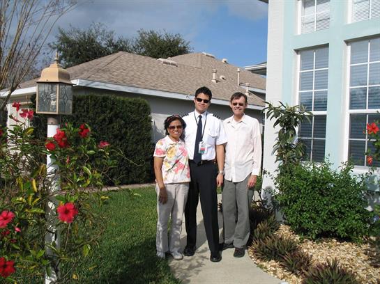 Two adults and a young man stand together in front of a house amidst blooming plants.