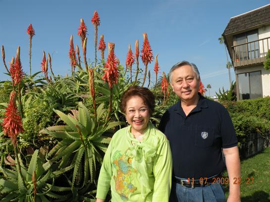 Older couple smiles broadly while standing among colorful flowers in a beautiful garden.