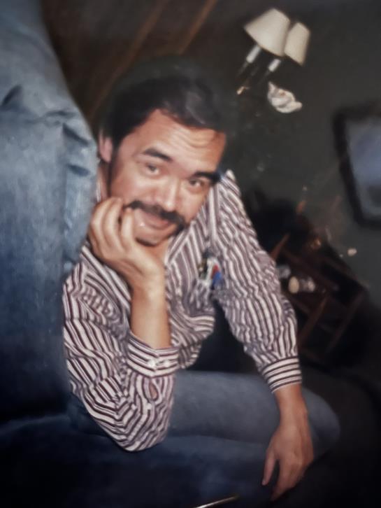 Man with a mustache sits comfortably in a relaxed pose, exuding a friendly vibe in his surroundings.