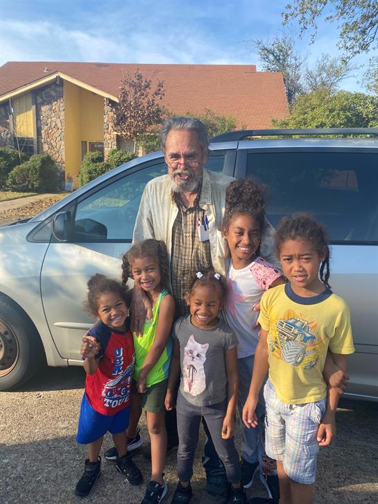 A group of children joyfully poses with a smiling man near a silver van on a bright day.