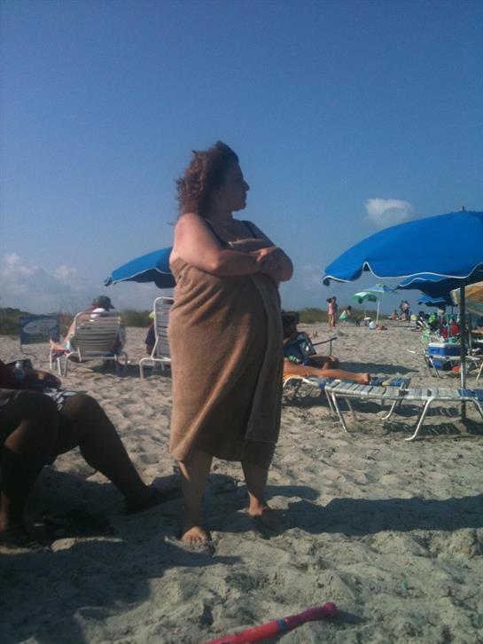 Woman with curly hair wraps a towel around herself while standing on the sandy beach.