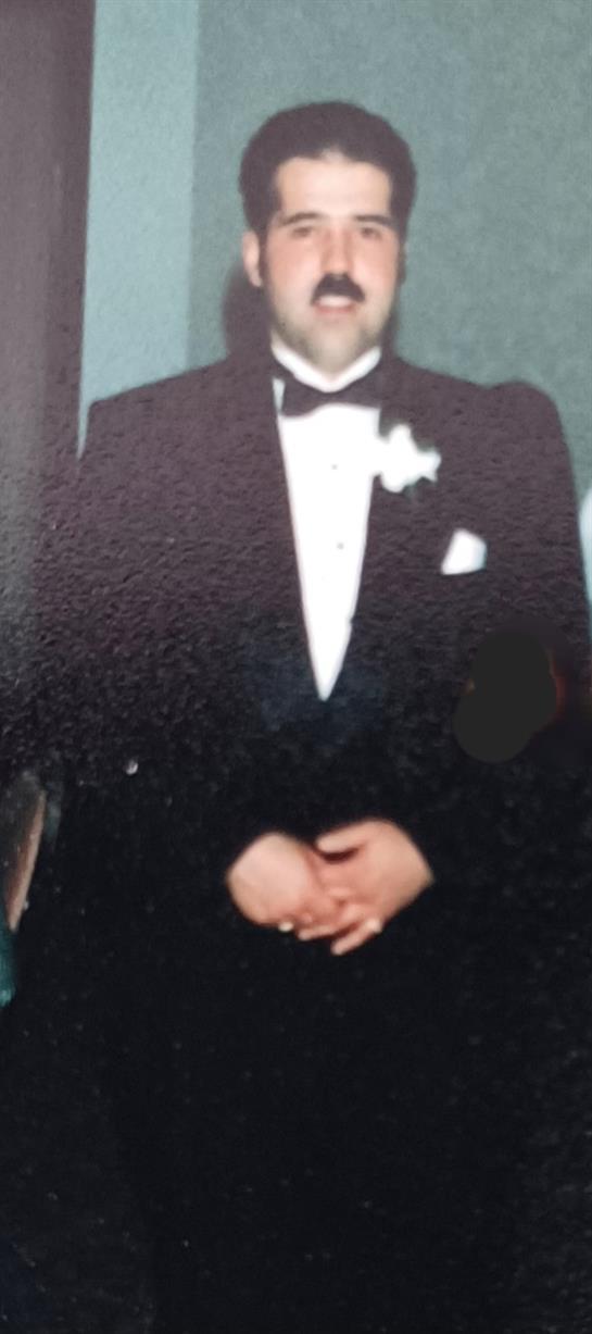 A well-dressed man in a tuxedo stands confidently, hands clasped, at an elegant event.